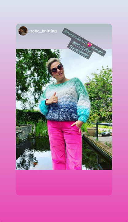 Person wearing a colorful sweater and pink pants standing by a pond with trees in the background.